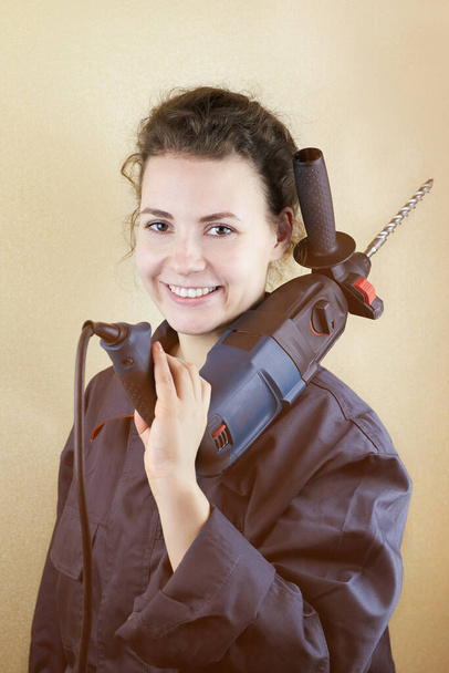 Χαμογελώντας όμορφη γυναίκα σε workwear με ένα puncher κοντά στον τοίχο, τονισμένη φωτογραφία. - Φωτογραφία, εικόνα