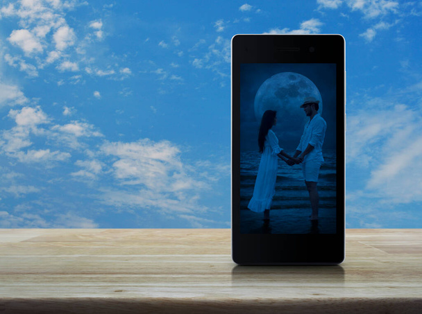 Romantic loving couple standing and holding hands together on modern smart mobile phone screen on wooden table over blue sky with white clouds, Business internet dating online, Valentines day concept - Foto, Imagem