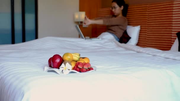 Фрукти в таці на ліжку в номері розкішного готелю перед азіаткою жінка подорожній розтягується і використовує ноутбук.. - Кадри, відео