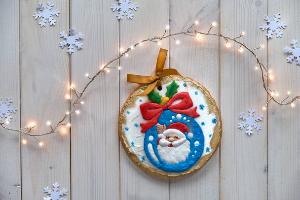 Decorated gingerbread. Cartoon Santa character on bauble with bow and holly. Creative traditional gifts for children. Christmas flat lay with snowflakes, garland of lights. Rustic off white wood table. - Photo, Image