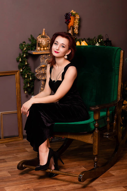 A young woman in a black dress sits in a rocking chair next to the fireplace and a Christmas tree. - Fotografie, Obrázek