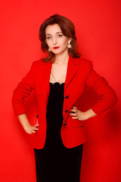 Attraktive junge Frau in schwarzem Kleid und roter Jacke posiert vor rotem Hintergrund. - Foto, Bild
