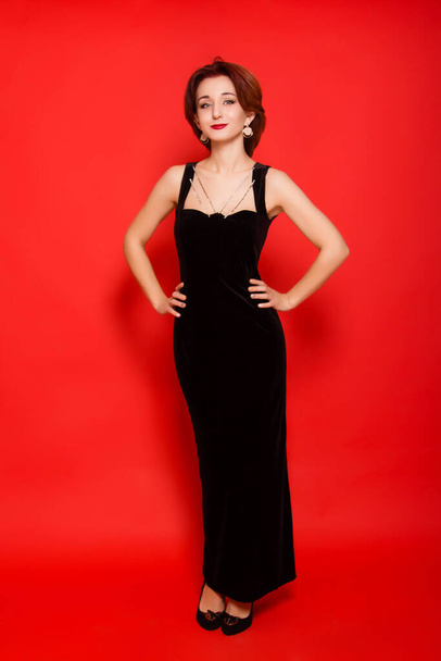 Beautiful, young woman in a black dress and black shoes posing standing on a red background. - Foto, afbeelding