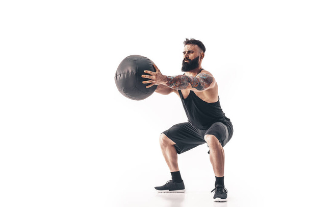 Muscular tattooed bearded male exercising fitness weights Medicine Ball in studio isolated on white background - Foto, afbeelding