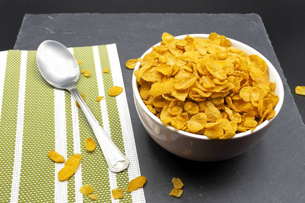 Een kom cornflakes als ontbijt naast een metalen lepel en een groene doek op een leisteenachtergrond - Foto, afbeelding