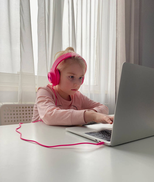 Vzdělání na dálku. Školačka v růžových sluchátkách studující domácí úkoly během své online lekce doma přes internet. Společenská vzdálenost během karantény - Fotografie, Obrázek
