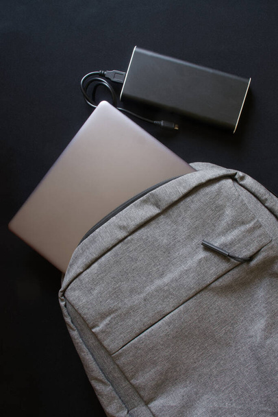 A gray textile city backpack with a silver laptop or ultrabook and an external battery on a black background. Traveling with gadgets. Black gradient - Фото, зображення