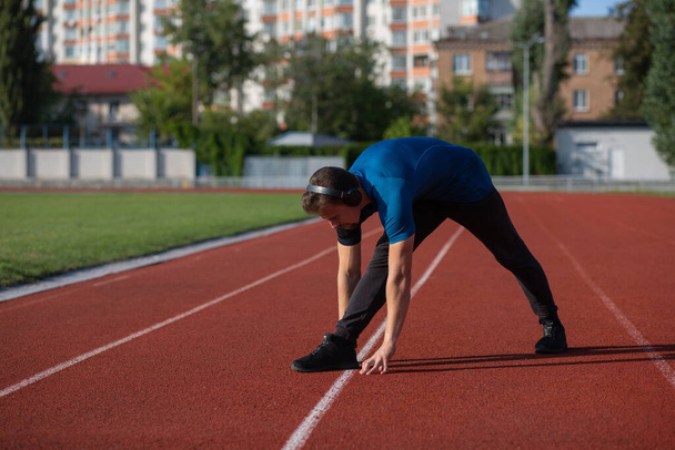 Ajuste joven hombre lleva auriculares haciendo estiramiento en la pista de atletismo. Espacio vacío - Foto, Imagen