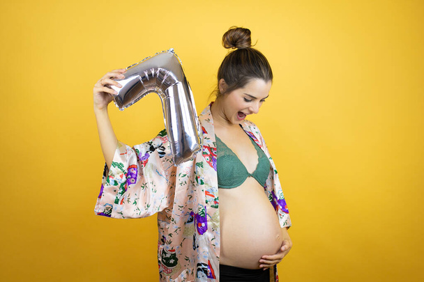 Jovem mulher bonita grávida esperando bebê vestindo pijama sobre fundo amarelo isolado segurando um número sete balão. Conceito de celebração - Foto, Imagem