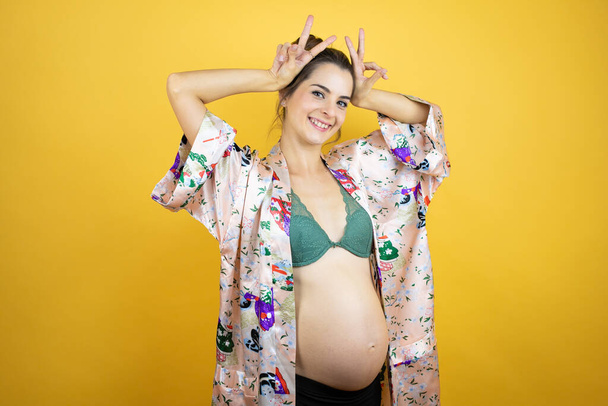 Молодая красивая женщина беременная ожидает ребенка в пижаме на изолированном желтом фоне Представляя смешно и безумно с пальцами на голове, как кролик уши, улыбаясь весело - Фото, изображение