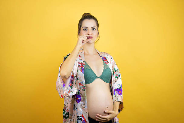 孤立した黄色い背景口の上にパジャマを被った赤ちゃんを期待して妊娠中の若い美少女と唇が指でzipとしてシャットダウンします.秘密裏に黙ってタブーを語る - 写真・画像