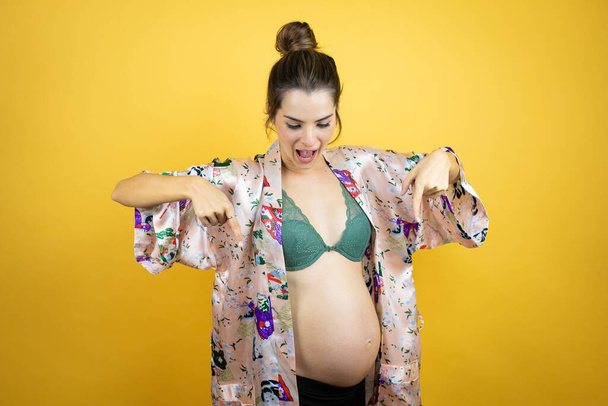 Fiatal gyönyörű nő terhes vár baba visel pizsama felett elszigetelt sárga háttér meglepett, nézett le, és rámutatva le ujjakkal és emelt karokkal - Fotó, kép