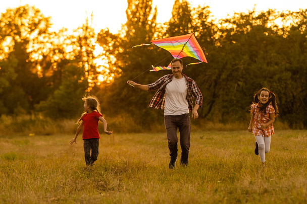 Ευτυχισμένη οικογένεια πατέρας και παιδί κόρη τρέχουν με έναν χαρταετό στο λιβάδι - Φωτογραφία, εικόνα