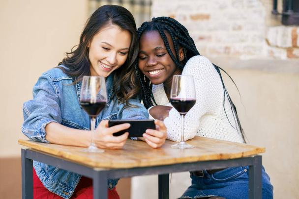 Twee vrouwen die samen naar hun smartphone kijken terwijl ze een glas wijn drinken. - Foto, afbeelding