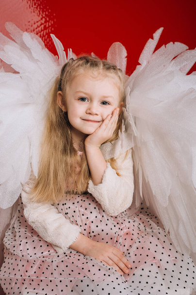 κοριτσάκι 4 ετών σε λευκό φτερά αγγέλου σε φόντο κόκκινο λάτεξ στο στούντιο για την ημέρα του Αγίου Βαλεντίνου - Φωτογραφία, εικόνα