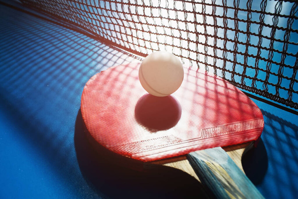 Una raqueta de tenis de mesa roja y una bola blanca yacen en la superficie de la mesa junto a la red. Deportes juego  - Foto, Imagen
