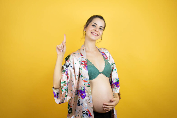 Jovem mulher bonita grávida esperando bebê vestindo pijama sobre fundo amarelo isolado mostrando e apontando para cima com os dedos número um enquanto sorri confiante e feliz - Foto, Imagem