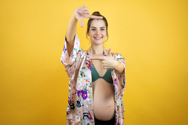 Young beautiful woman pregnant expecting baby wearing pajama over isolated yellow background smiling making frame with hands and fingers with happy face - 写真・画像