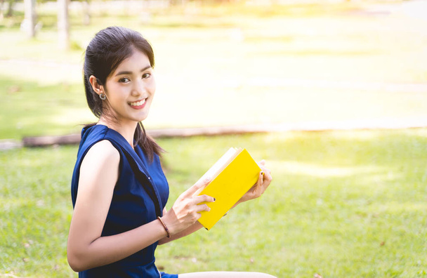 Ασιάτισσες διαβάζουν ένα κίτρινο βιβλίο ή μυθιστόρημα, απολαμβάνοντας και χαρούμενος, η γυναίκα ανοίγει ένα βιβλίο στο χέρι και κατά λάθος να το διαβάσετε στο πάρκο θολό φόντο με αντίγραφο χώρο - Φωτογραφία, εικόνα