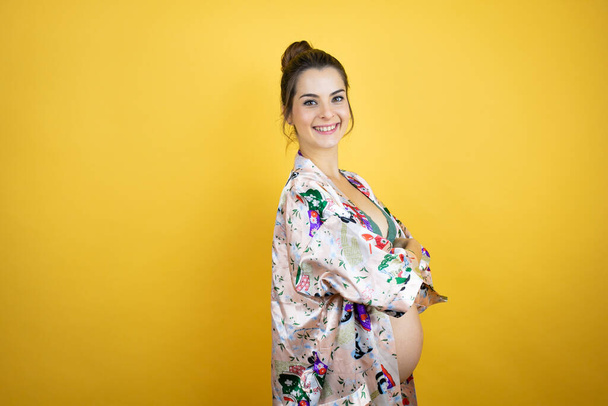 Junge schöne schwangere Frau, die ein Baby erwartet, trägt Pyjama über isoliertem gelbem Hintergrund und schaut zur Seite, die Arme verschränkt und überzeugt - Foto, Bild