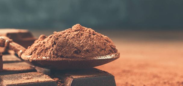 Коричневый какао порошок в ложке и нарезанные кубики шоколада, на темном фоне с копировальным местом. - Фото, изображение