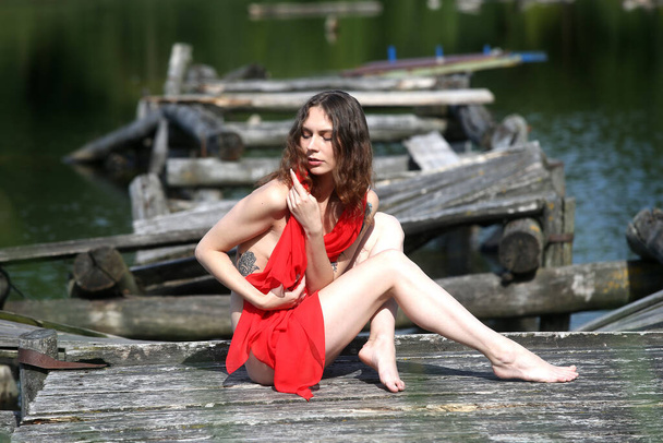 Kaunis alaston tyttö, jolla on upeat jalat punaisessa materiaalissa, poseeraa vanhalla puisella sillalla  - Valokuva, kuva
