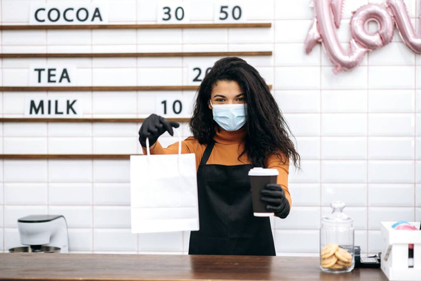 Jeune fille barista afro-américaine en uniforme et masque médical et gants de protection donne un café de visiteur dans un verre en carton jetable et dessert dans un sac en papier à emporter. Café à emporter concept. - Photo, image