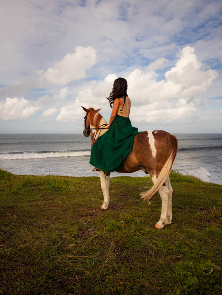 Žena jezdící na koni u oceánu. Venkovní aktivity. Asijka v dlouhých zelených šatech. Koncept cestování. Zataženo. Pohled zezadu. Vertikální uspořádání. Rozumím. Bali, Indonésie - Fotografie, Obrázek