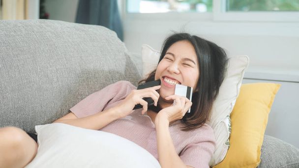 Young smiling Asian woman using smartphone buying online shopping by credit card while lying on sofa when relax in living room at home. Lifestyle latin and hispanic ethnicity women at house concept. - Photo, Image