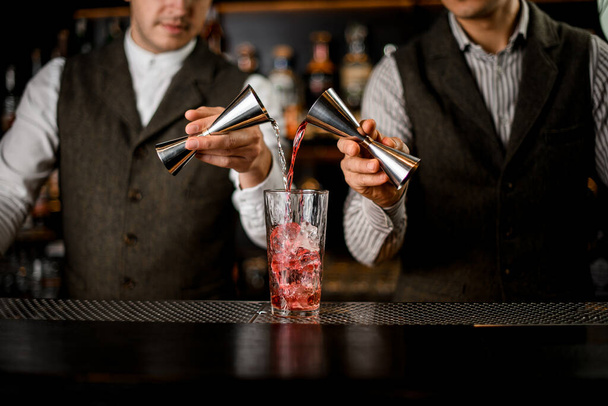 bartenders hold steel jiggers and pour drink into glass with ice cubes - Φωτογραφία, εικόνα