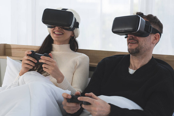 couple caucasien assis dans le lit dans la chambre à coucher jouer jeu de réalité virtuelle ensemble à la maison - Photo, image