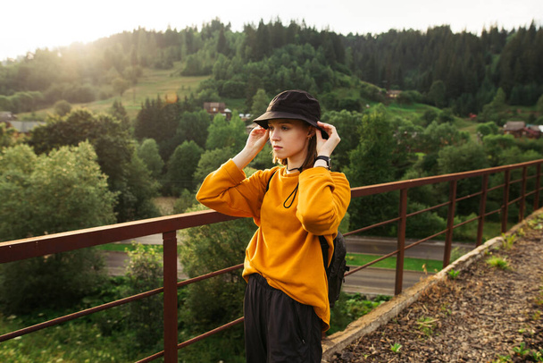 Portré egy vonzó női túrázó narancssárga pulóver és kalap áll egy hídon a hegyekben, és néz félre egy komoly arc. - Fotó, kép