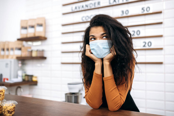 Üniformalı ve koruyucu tıbbi maske takan üzgün, genç, Afro-Amerikan bayan kahve dükkanı tezgahının arkasında müşterileri bekliyor. Kahve barı konsepti - Fotoğraf, Görsel