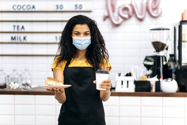Young african american waitress girl in uniform and medical mask gives a visitor coffee in a disposable cardboard glass and dessert. Service industry concept, coffee shop - Photo, Image