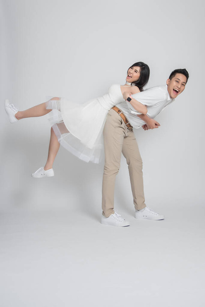 Happy Asian groom gives a bride piggyback ride having fun on white background. - Φωτογραφία, εικόνα