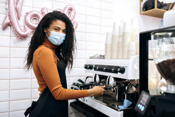 Молодая афроамериканка-бариста в медицинской маске и черном фартуке готовит кофе для посетителей кофейни. Владелец малого бизнеса. Кафе, концепция сферы услуг - Фото, изображение