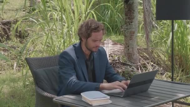 Średni strzał białego przystojnego biznesmena pracującego na laptopie siedzącego przy biurku pod palmami w słoneczny dzień lata - Materiał filmowy, wideo
