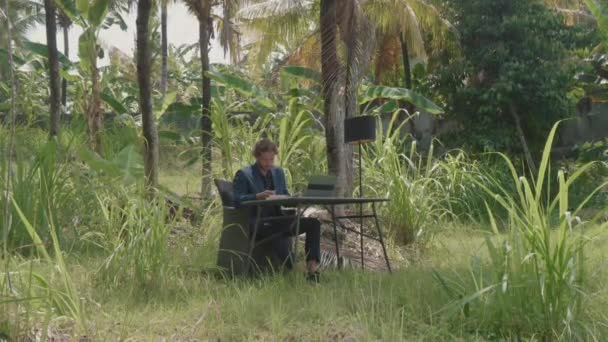 Široký záběr pohledného obchodně vypadajícího muže sedícího za stolem pod palmami venku na bali venkově a pracujícího na notebooku za denního světla - Záběry, video