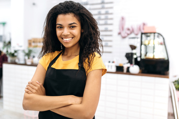 Aantrekkelijke jonge Afro-Amerikaanse vrouwelijke barista met krullend haar in zwart schort staat met gekruiste armen in cafe en vriendelijke glimlach. Portret van een mooie serveerster in uniform - Foto, afbeelding