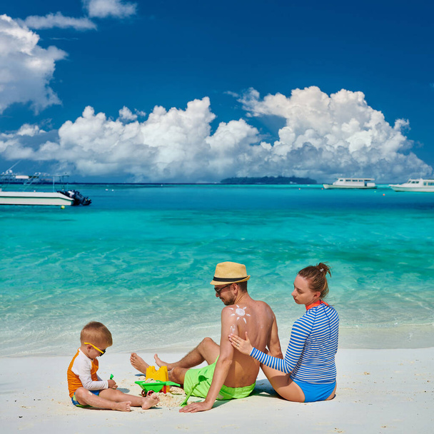 Rodina na pláži, mladý pár s tříletým chlapcem. Žena nanáší opalovací krém na pánská záda. Letní dovolená na Maledivách. - Fotografie, Obrázek