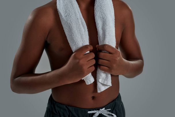 Cropped shot of a half naked african boy holding towel on shoulders, resting after workout while standing against grey background - Foto, Imagem