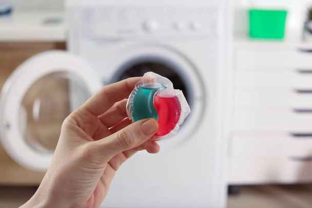 Woman holding laundry detergent capsule near washing machine indoors, closeup - Fotoğraf, Görsel