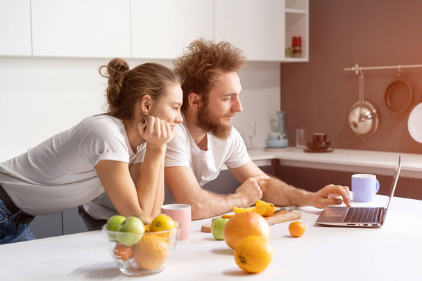 Leaned on kitchen table couple watching romantic movie or streaming social media video. Beautiful young couple talking on video call using laptop. Young couple in kitchen at new home - Foto, imagen