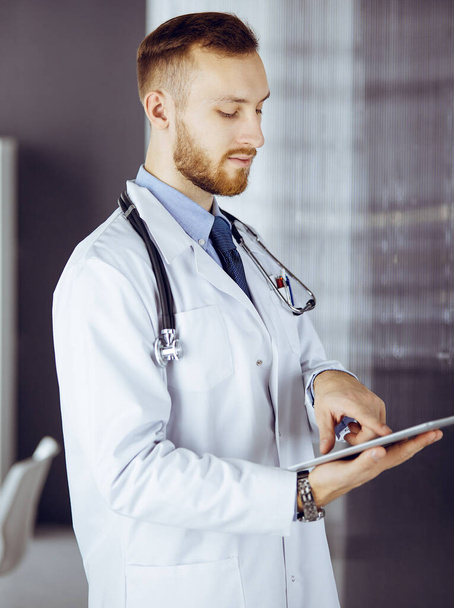 Friendly red-bearded doctor using tablet computer in clinic at his working place. Perfect medical service in hospital and medicine concept - Photo, image