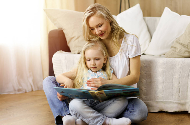 Happy family. Blonde young mother reading a book to her cute daughter while sitting at wooden floor in sunny room. Motherhood concept - Фото, зображення
