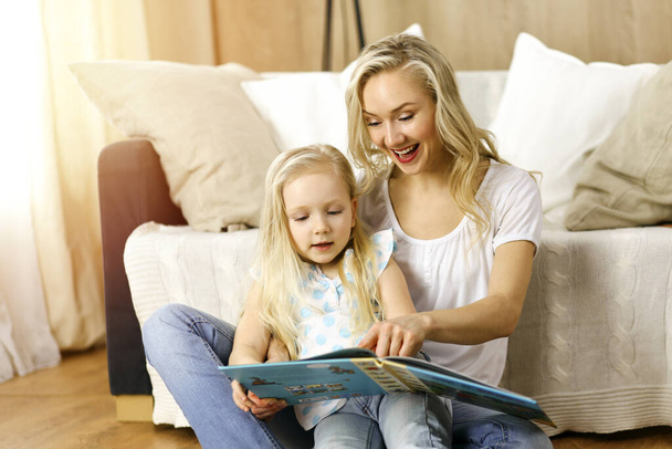 Happy family. Blonde young mother reading a book to her cute daughter while sitting at wooden floor in sunny room. Motherhood concept - Fotografie, Obrázek