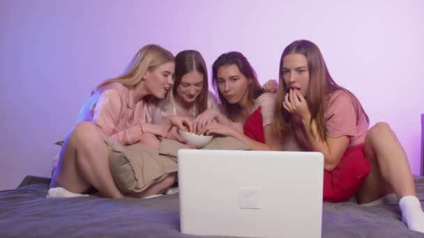 Quatre jolies jolies jeunes femmes en pyjama s'assoient sur le lit, mangent du pop-corn et regardent un film au enterrement de vie de jeune fille - Séquence, vidéo