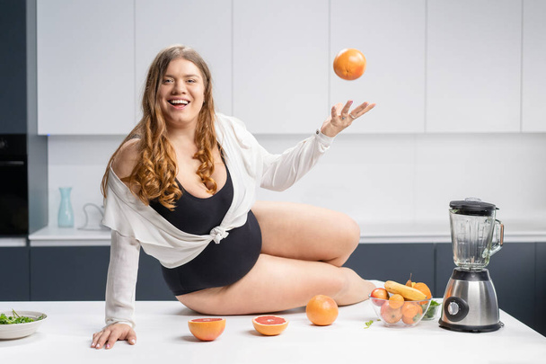 Girl with obese problem. Young sexy chubby white girl in black swimsuit, white shirt at modern kitchen table. juggling fruits in hand. Trying to loose weight fast. Fat barefoot girl low carb diet - Φωτογραφία, εικόνα