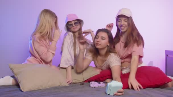 Τέσσερις χαρούμενες όμορφες γυναίκες με πιτζάμες κάθονται στο κρεβάτι και παίρνουν μια μεταξωτή φωτογραφία σε vintage κάμερα με γυαλιά ηλίου και καπέλα στο μπάτσελορ πάρτι - Πλάνα, βίντεο