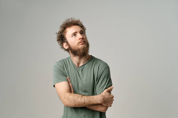 Portrait of pensive deep in thoughts young man with hands folded. Attractive man with curly hair and beard in olive t-shirt looking at camera isolated on white background - 写真・画像